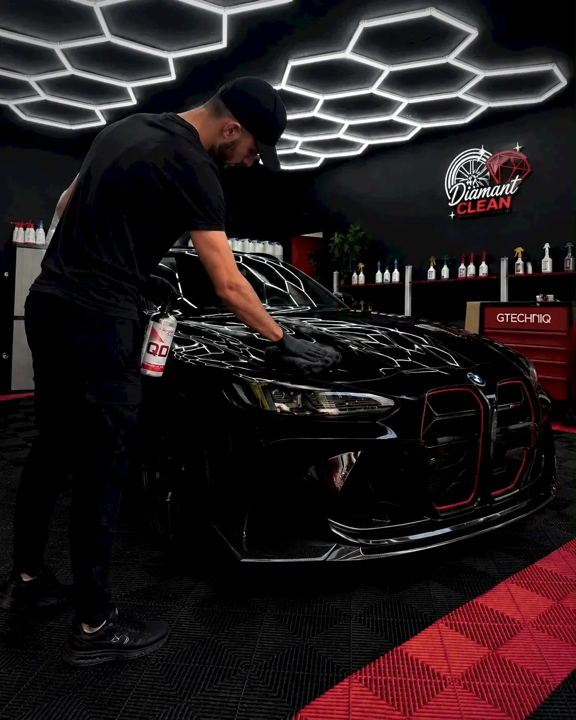 Préparateur DiamantClean essuyant le capot d'une BMW noire dans l'atelier, logo DiamantClean au mur